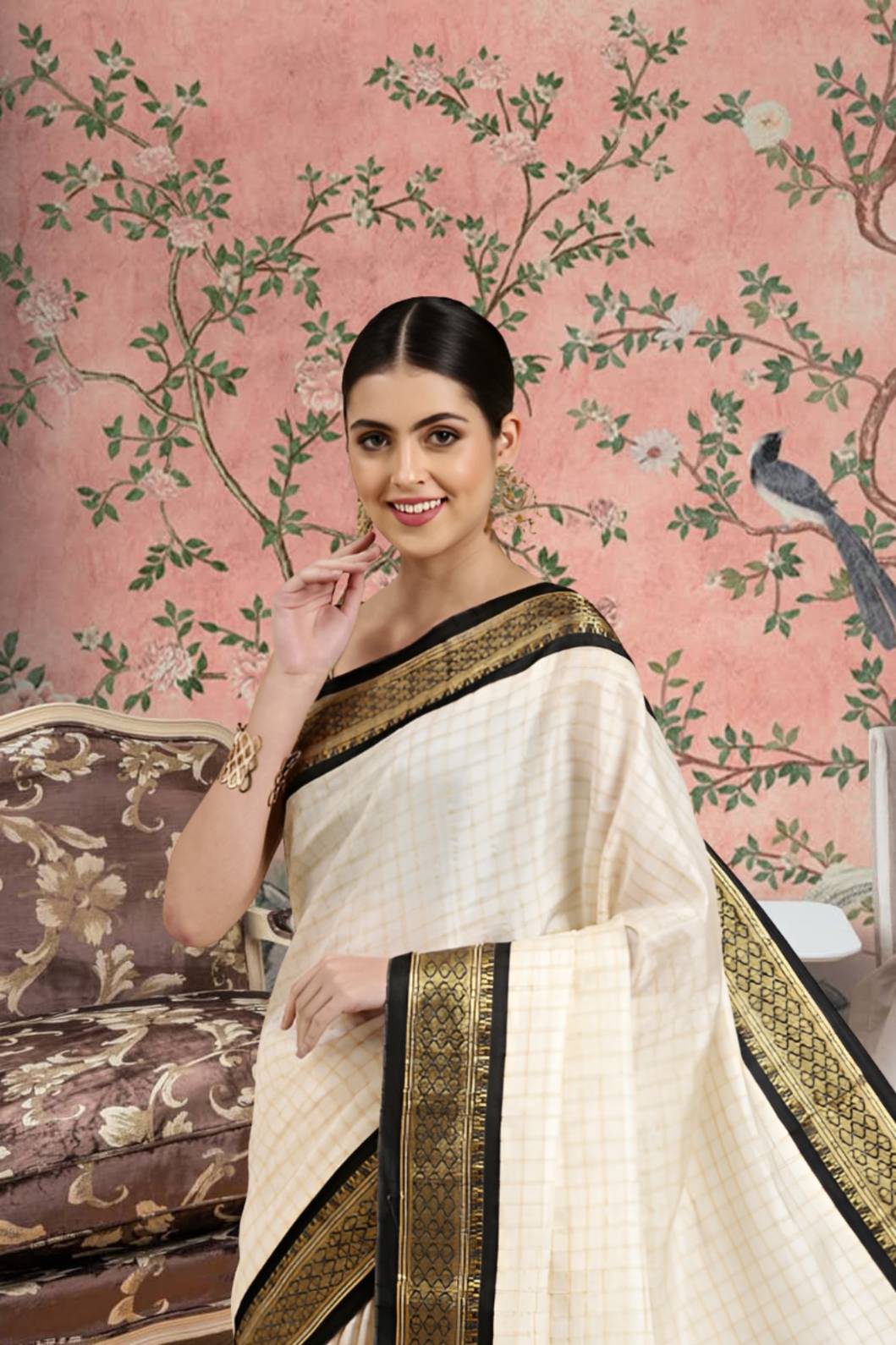 Woman wearing a white saree with black and gold border against a decorative pink wall.
