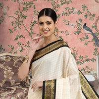 Woman wearing a white saree with black and gold border against a decorative pink wall.