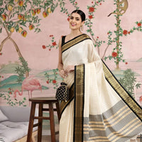 Woman in a white saree with black and gold border standing in a room with decorative wall art.