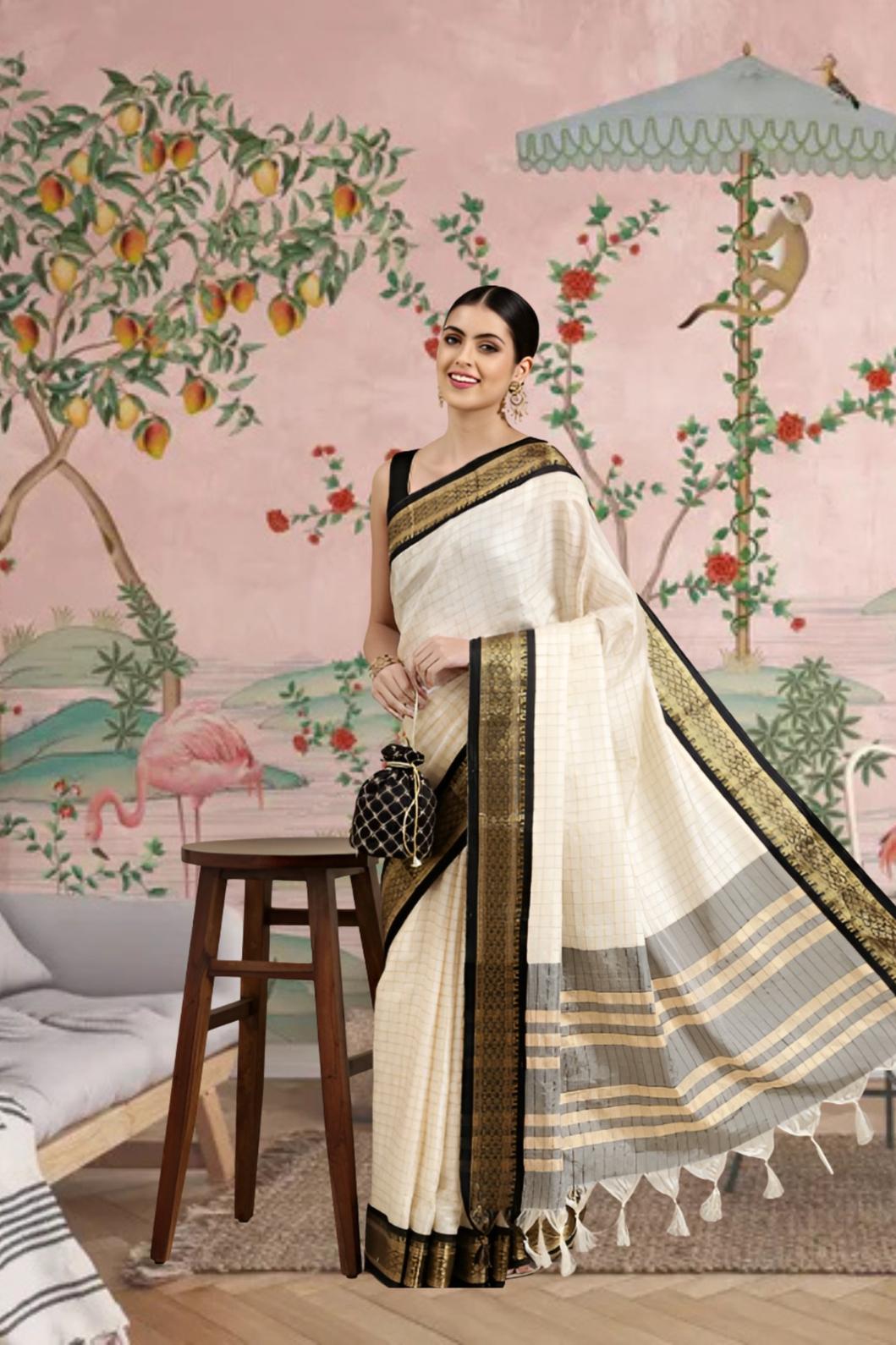 Woman in a white saree with black and gold border standing in a room with decorative wall art.