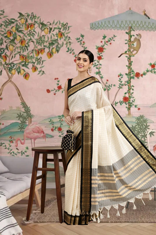 Woman in a white saree with black and gold border standing in a room with decorative wall art.